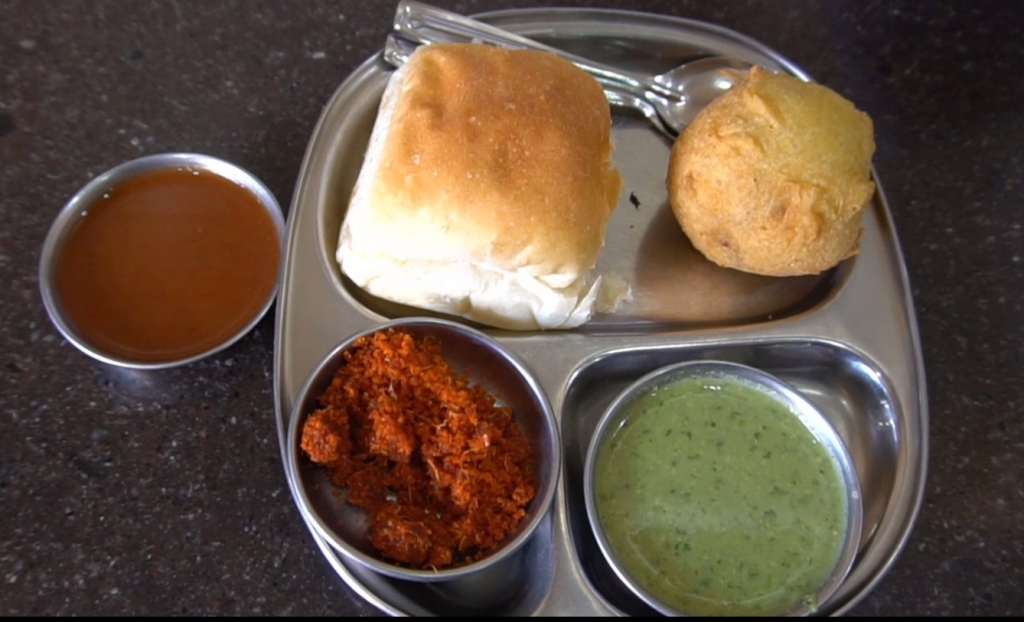 A plate of vada pav served along with dry garlic chutney and runny coconut mint and tamarind chutneys,